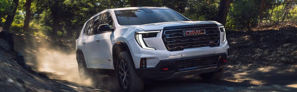 White GMC SUV driving off-road through a dusty forest trail surrounded by trees.