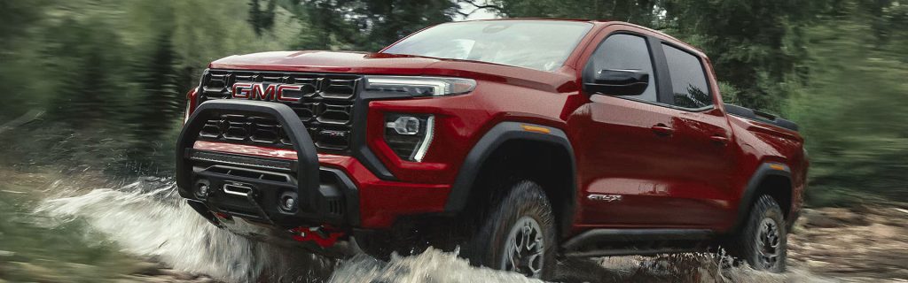 Red GMC pickup truck splashing through a shallow stream on a forest trail